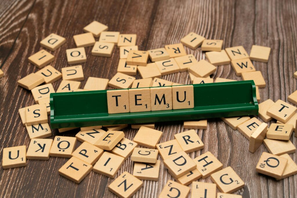 Wooden Scrabble tiles spelling TEMU on a green rack with scattered tiles on a wooden surface. Scattered letters on a desk forming the word Temu, symbolizing the Temu phenomenon in e-commerce.