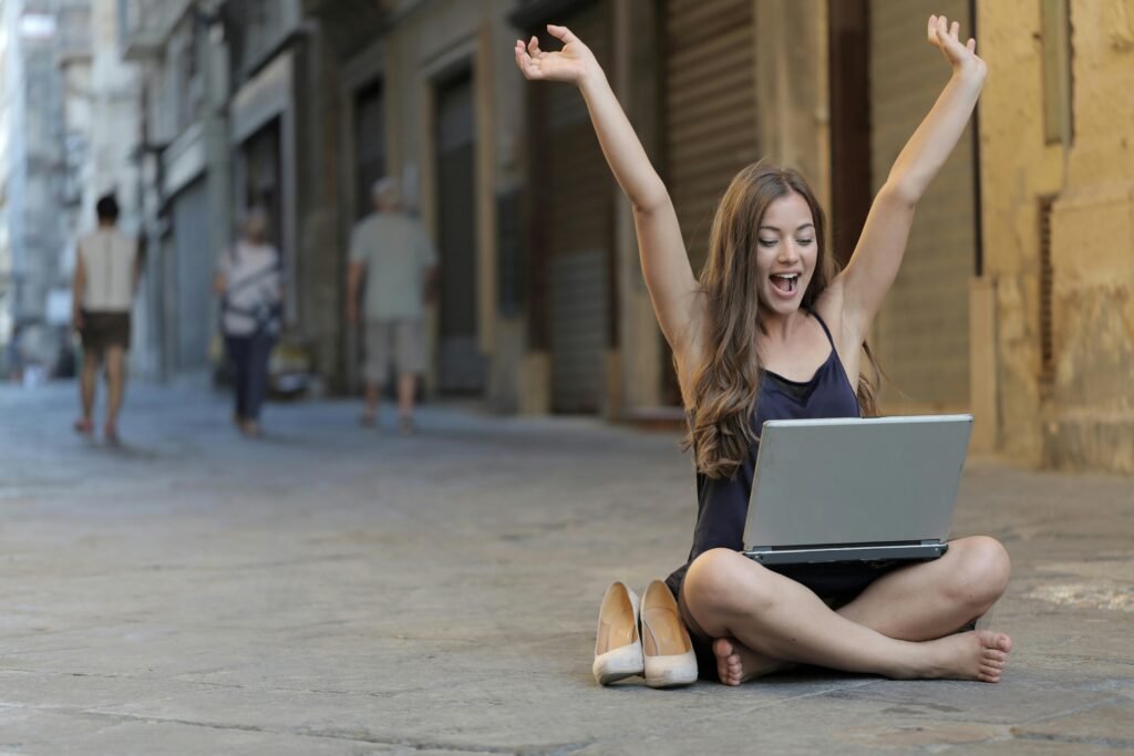pexels-photo-3813341-3813341 A cheerful woman sitting outdoors, celebrating success with arms raised, while using a laptop.